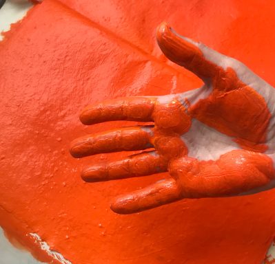 Aufsicht auf eine Handinnenseite und Finger, die mit oranger Farbe bedeckt sind, im Hintergrund oranges Papier.